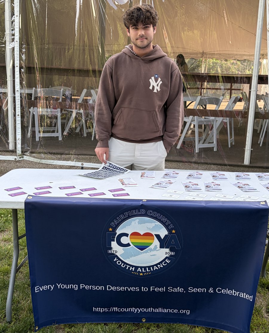 Milo Mutuski, founder of the Fairfield County LGBTQ+ Youth Alliance, standing behind the organization's table at a community event.